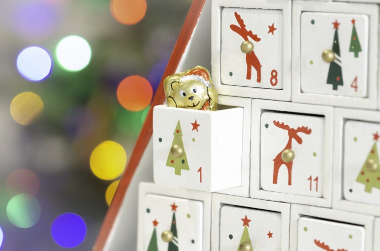 A close-up of a Christmas advent calendar with small white drawers. One drawer is open, revealing a gold foil-wrapped chocolate bear. The drawers are decorated with Christmas-themed images like trees and reindeer. Colorful lights are blurred in the background.