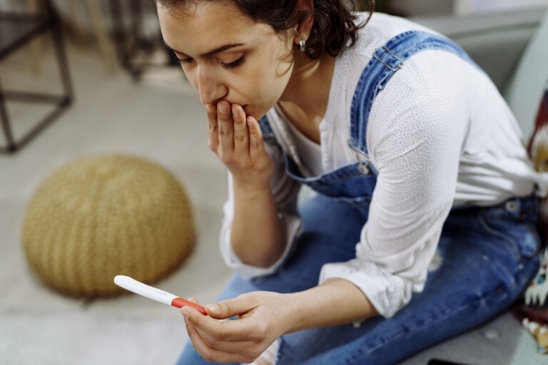 A woman sitting on a couch, wearing blue overalls and a white shirt, holds a pregnancy test in one hand while covering her mouth with the other, looking intently at the result—pondering if it's possible for a stealth pregnancy. A round woven pouf is in the background.