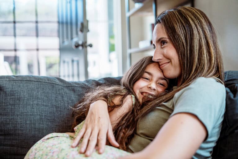 A woman with long hair and a grey shirt sits on a couch, embracing a young girl with long hair. The girl, smiling and resting her head on the womans chest, seems unaware of what causes a child to be disrespectful. An open door and bookshelf are seen in the background.