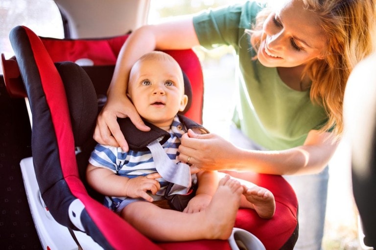A woman secures a fussy baby into a car seat inside a vehicle. The child is seated in a red and black car seat, while the woman adjusts the straps. The baby, wearing a blue and white striped shirt, looks upwards with an expression of mild discomfort.