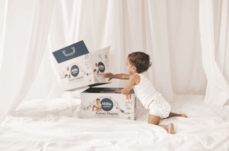 A toddler wearing a white tank top and diaper is kneeling on a white surface, reaching into an open box of Millie Moon Luxury Diapers. Another box of these premium diapers is visible beside it. The background consists of white curtained drapes.